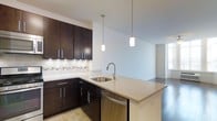 This single-floor residence showcases a thoughtfully designed open-concept layout that connects the kitchen and living areas in a seamless, functional flow. The kitchen is a standout feature, appointed with rich dark espresso cabinetry equipped with sleek brushed nickel hardware. Light granite countertops provide a striking contrast against the dark cabinetry, while a mosaic tile backsplash adds a refined decorative touch. A peninsula with an integrated stainless steel sink extends the counter space and doubles as a casual dining or prep area. Two elegant cylindrical pendant lights hang above the peninsula, casting warm, focused illumination. Stainless steel appliances, including a dishwasher, complement the modern aesthetic. The kitchen transitions naturally into the adjoining living space, which features dark hardwood flooring and bright white walls that create a clean, contemporary atmosphere. Natural light filters in through a window visible in the background, enhancing the open and airy feel of the space. The combination of modern finishes, quality materials, and a well-considered layout makes this a highly functional and visually appealing living environment.