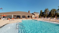 This pool amenity space combines an inviting outdoor environment with a well-appointed interior clubhouse, creating a comprehensive recreational retreat. Outside, a generously sized swimming pool takes center stage, framed by a spacious sundeck lined with lounge chairs in vibrant orange accents. The adjoining clubhouse building features a classic roofline with brick detailing and large glass doors that seamlessly connect the indoor and outdoor areas.

Inside, the clubhouse is designed with modern sophistication. Polished concrete floors run throughout, lending a sleek, contemporary feel to the space. The main gathering area boasts soaring vaulted ceilings with large ceiling fans and recessed lighting, anchored by a stone-clad fireplace with a wall-mounted television above. Comfortable lounge seating and a media console create a relaxed social atmosphere.

A dining area features a round wooden table with leather chairs, positioned near floor-to-ceiling glass doors that offer direct views of the pool. A bar area with dark granite countertops, warm wood cabinetry, a mosaic tile backsplash, and industrial-style pendant lights provides an ideal spot for casual entertaining. The entryway is accented with contemporary artwork and warm-toned furnishings, completing the refined and welcoming character of this amenity space.