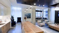 This contemporary single-floor space showcases a thoughtfully designed interior that blends industrial and modern aesthetics seamlessly. A striking dark ceiling contrasts with bright white walls and polished concrete floors, creating a sophisticated visual balance throughout. Exposed concrete columns serve as architectural focal points, reinforcing the space's urban, industrial character.

At the center, a substantial live-edge wooden communal table anchors the room, offering a warm, natural counterpoint to the sleek surroundings. Overhead, a cluster of globe pendant lights with mirrored finishes adds a refined, decorative touch while providing ambient illumination.

A relaxed lounge area features dark, high-backed armchairs arranged to encourage comfortable conversation, while a separate seating zone with neutral-toned chairs and round tables provides a more casual setting. Toward the perimeter, floor-to-ceiling windows flood the space with generous natural light and offer open views of the surrounding urban environment. Bar-height seating lines the window wall, creating an ideal spot for work or informal gatherings.

Curated artwork adorns the walls, contributing to the space's polished yet approachable atmosphere. Overall, the layout is versatile and well-suited for collaboration, socializing, or focused individual use.