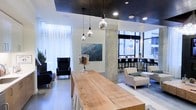 This contemporary single-floor space showcases a thoughtfully designed interior that blends industrial and modern aesthetics seamlessly. A striking dark ceiling contrasts with bright white walls and polished concrete floors, creating a sophisticated visual balance throughout. Exposed concrete columns serve as architectural focal points, reinforcing the space's urban, industrial character.

At the center, a substantial live-edge wooden communal table anchors the room, offering a warm, natural counterpoint to the sleek surroundings. Overhead, a cluster of globe pendant lights with mirrored finishes adds a refined, decorative touch while providing ambient illumination.

A relaxed lounge area features dark, high-backed armchairs arranged to encourage comfortable conversation, while a separate seating zone with neutral-toned chairs and round tables provides a more casual setting. Toward the perimeter, floor-to-ceiling windows flood the space with generous natural light and offer open views of the surrounding urban environment. Bar-height seating lines the window wall, creating an ideal spot for work or informal gatherings.

Curated artwork adorns the walls, contributing to the space's polished yet approachable atmosphere. Overall, the layout is versatile and well-suited for collaboration, socializing, or focused individual use.