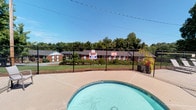 The Kids Pool area is an outdoor recreational space designed with younger swimmers in mind. The centerpiece is a compact, rounded shallow pool with clear blue-green water, purpose-built for safe use by children. The pool is enclosed within a chain-link fence perimeter, providing a secure boundary and clear sightlines for supervision. Posted signage, including no-diving notices, reinforces the safety-focused nature of the space.

The surrounding concrete pool deck offers ample open space for movement and relaxation. A shaded patio area adjacent to the pool features a white outdoor dining table with seating for four and a large umbrella overhead, providing a comfortable spot for supervising adults or those taking a break from the water. Potted plants and a low brick planter add a touch of greenery to the patio setting.

A small brick utility building is visible nearby, and a gated entry point allows controlled access to the fenced area. The broader surroundings include mature trees that frame the space with natural shade and privacy. Views from the pool deck reveal an adjacent larger pool area with lounge chairs, indicating this kids pool is part of a wider aquatic amenity complex. The open sky and lush tree canopy create a bright, welcoming outdoor atmosphere.