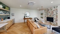 Located at 4729 Manzanita Ave in Carmichael, CA, this single-floor combined living and dining space offers a thoughtfully designed interior with a clean, modern aesthetic. The entire space is unified by striking herringbone-patterned hardwood flooring that adds warmth and visual texture throughout.

The dining area features a sleek dark wood dining table paired with woven-strap chairs and is anchored by a light natural wood credenza topped with curated décor. Framed abstract artwork in teal and earth tones adorns the crisp white walls. Large windows and a glass-panel door flood the area with natural light and provide views of the lush greenery outside.

The living area transitions seamlessly from the dining space and centers around a tufted tan leather sofa, complemented by a woven rattan pendant light and a round mirror above a mid-century console. A dramatic botanical-print accent wall adds a bold design moment near the sitting area.

A built-in wet bar or refreshment station serves as a functional and stylish highlight, featuring deep green subway tile that extends to the ceiling, open shelving, and warm wood cabinetry — creating a rich, spa-like focal point. Recessed lighting and a cassette-style ceiling HVAC unit ensure year-round comfort. The overall ambiance is bright, curated, and inviting.