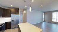 This single-floor space showcases a clean, modern design with an open-concept layout that seamlessly connects the kitchen and living areas. The kitchen is outfitted with sleek, dark wood cabinetry offering ample storage, a stainless steel refrigerator with an integrated ice and water dispenser, and a large white countertop island that serves as both a functional workspace and a casual gathering point. Two cylindrical pendant lights hang above the island, providing focused task lighting while adding a contemporary design element. Recessed lighting throughout the space ensures consistent, well-distributed illumination. The light grey walls and light hardwood-style flooring contribute to a bright, neutral palette that enhances the sense of openness throughout the space. A hallway visible from the kitchen leads further into the unit, suggesting additional rooms beyond the main living area. Natural light filters in through a window near the living area, adding warmth to the overall ambiance. The combination of modern fixtures, neutral tones, and an open floor plan creates a functional yet stylish environment suited for everyday living.