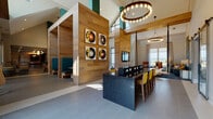 Located at 330 Pennington Centre Blvd in Nashville, TN, this single-floor community clubhouse is a thoughtfully designed gathering space that draws inspiration from Nashville's rich musical heritage. The open-concept layout is anchored by soaring ceilings with dramatic height, creating an airy, expansive atmosphere throughout.

The main lounge area features large floor-to-ceiling windows that flood the space with natural light and offer open views of the surrounding landscape. Comfortable seating — including warm leather armchairs, plush sofas, and bar-height tables with teal and yellow accent chairs — is arranged throughout the space to encourage both social interaction and relaxed lounging.

A standout feature is the wood-paneled interior wall structure housing intimate teal-upholstered booth seating, adorned with bold vinyl record artwork that pays homage to Nashville's musical identity. A striking circular chandelier with exposed Edison bulbs serves as a focal point overhead.

The kitchen area includes a large white quartz island with patterned tile flooring, pendant lighting, and a built-in wet bar with a glass garage-style door that opens to the outdoors. A game room with a pool table and additional lounge seating rounds out the space. Throughout, the design blends warm wood tones, polished tile floors, and vibrant pops of color for a lively yet sophisticated ambiance.