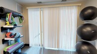 This single-floor space is defined by a prominent sliding glass door fitted with full-length white vertical blinds that span the entire width of the wall. The blinds provide effective light control while allowing diffused natural light to filter softly into the room, creating a bright and airy atmosphere. The walls are finished in a warm neutral tone, contributing to an inviting and understated aesthetic throughout the space.

Ceiling-mounted HVAC vents are positioned above the sliding door, indicating a centralized climate control system. A wall-mounted air conditioning unit is also visible along the upper portion of one wall, offering supplemental temperature management. The flooring appears to be a smooth tile, which complements the clean, practical character of the room.

Along one wall, open shelving provides functional storage and display space, adding a modest organizational element to the room. The overall layout is straightforward and uncluttered, with a design emphasis on practicality and ease of use. The combination of natural light access through the glass door, neutral finishes, and efficient climate control makes this a versatile and functional living area.