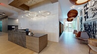 This elegantly designed lobby serves as the welcoming hub of a modern residential building, thoughtfully composed to balance functionality with refined aesthetics. The reception area anchors the entrance, featuring a wood-clad desk with a dark stone countertop and an eye-catching sculptural ceiling installation of layered white pendant lights. Oversized warm-toned orb pendants line the main corridor, drawing the eye toward glass entry doors that flood the space with natural light and offer views of lush greenery beyond.

The flooring transitions seamlessly between large-format light stone tiles and wide-plank white oak hardwood, unifying the distinct zones of the lobby. A standout lounge area is defined by soaring ceilings, a bold elongated linear fireplace set into a dark feature wall, and a curated arrangement of velvet and leather seating atop a richly colored abstract area rug. Designer tiered chandelier fixtures illuminate this space with warmth and drama.

Additional highlights include a glass-enclosed meeting or amenity room with wood and mirror accents, a living green wall installation, and a dedicated elevator bank clad in reclaimed wood planking. Throughout, the space projects a contemporary, upscale character through its careful layering of natural materials, curated artwork, and statement lighting.