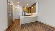 This contemporary single-floor residence is thoughtfully designed with a clean, modern aesthetic and high-quality finishes throughout. Rich hardwood-style flooring flows seamlessly across the main living and kitchen areas, creating a warm and cohesive feel.

The open-concept living area is bright and airy, featuring crisp white walls, a ceiling fan, and a large window that frames views of the surrounding urban streetscape, allowing natural light to fill the space generously.

The kitchen is a standout feature, equipped with custom shaker-style cabinetry in a warm taupe finish, sleek white quartz countertops, and premium stainless steel appliances including a double oven range with a wall-mounted hood and a French door refrigerator. A peninsula with a waterfall countertop edge provides additional prep space and casual seating, while open shelving adds a stylish, functional touch.

The primary bathroom offers a dual-sink vanity with undermount basins, quartz countertops, and floor-to-ceiling cabinetry providing ample storage. A secondary bathroom features a clean, minimalist design with wood-look tile flooring and a single vanity. The carpeted bedroom is bathed in natural light through a trio of large windows overlooking the neighborhood exterior.