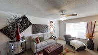 This single-floor space centers around a welcoming living room that balances comfort and eclectic style. The room features a textured ceiling with a ceiling fan and light fixture that provides both airflow and ambient lighting. Natural light filters in through a window fitted with blinds, brightening the carpeted interior. The living area is anchored by a gray sofa accented with colorful throw pillows in earth tones, complemented by a tufted burgundy leather ottoman that doubles as a coffee table. A patterned accent chair adds a bold visual contrast. The walls are adorned with large mixed-media art panels in dark, metallic tones, alongside a pair of circular decorative mirrors that add depth and dimension. A sleek side table with a decorative lamp provides additional warmth. The diagonal wall angle on one side gives the room an architectural character, creating a dynamic layout. The neutral wall tones and carpet provide a versatile backdrop for the decorative elements, resulting in a space that feels curated yet livable. Overall, the room presents a layered, personalized aesthetic with a mix of textures and warm accent colors.