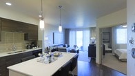 This contemporary single-floor residence showcases a thoughtfully designed open-concept floor plan that effortlessly unifies the kitchen, dining, and living spaces into one cohesive environment. The kitchen is anchored by a sleek white waterfall island with seating, illuminated by elegant cylindrical pendant lights that hang at varying heights, creating a warm and inviting focal point. Dark wood cabinetry lines the kitchen walls, offering a striking contrast against the light countertops, while modern appliances complete the functional workspace. The dining area flows naturally from the kitchen island, maintaining a seamless visual connection throughout. The living room is generously proportioned, featuring rich dark hardwood flooring and a neutral color palette accented by cool-toned furnishings. A large sofa arrangement centers the space, complemented by a sleek coffee table and a wall-mounted flat-screen television. Floor-to-ceiling vertical blinds frame a wide sliding glass door that bathes the interior in abundant natural light and hints at an outdoor access point beyond. The overall ambiance is polished and modern, with consistent finishes, recessed lighting, and carefully curated décor that lend the space a sophisticated yet comfortable character throughout.