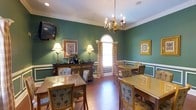This single-floor space exudes a traditional, refined ambiance through its carefully curated design elements. The room is finished with deep hunter-green walls accented by crisp white crown molding and a chair rail, lending it a distinguished, classic character. A central arched window with decorative drapery allows natural light to filter into the space, creating a welcoming focal point along the back wall. Overhead, a brass candelabra-style chandelier adds an elegant touch, complementing the warm-toned hardwood flooring that runs throughout. Multiple dining tables, each surrounded by upholstered chairs with a floral motif, are arranged to accommodate several guests comfortably, suggesting a communal dining or meeting function. A dark wood sideboard along one wall is dressed with table lamps and a floral arrangement, adding warmth and depth. Framed artwork adorns multiple walls, contributing to the room's polished, homey feel. A wall-mounted television provides a modern convenience within the otherwise traditionally styled setting. The overall atmosphere is warm, formal yet approachable, and well-suited for shared meals or social gatherings.