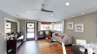 This single-floor space offers a thoughtfully designed interior that balances comfort and style. The main living area is the centerpiece of the space, featuring rich hardwood flooring that adds warmth and a polished look throughout. Neutral gray walls provide a clean, modern backdrop, complemented by warm terracotta and burnt orange accent tones that bring depth and character to the room.

The living area is generously proportioned, accommodating a plush light-toned sofa, coordinating accent chairs, and a sleek glass-top coffee table, all arranged on an earthy area rug that ties the seating together. A ceiling fan with a brushed nickel finish and recessed lighting provide both functionality and a contemporary aesthetic overhead.

Natural light pours in through multiple windows dressed with white curtains and horizontal blinds, creating a bright and airy atmosphere. A glass-paneled entry door further enhances the sense of openness and connectivity to the outdoors. Framed artwork on the walls adds a curated, finished feel to the space.

Overall, this space presents a warm, inviting ambiance with a clean and modern design sensibility, making it well-suited for both relaxed everyday use and welcoming guests.