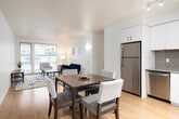 This well-appointed single-floor residence offers a thoughtfully designed open-concept layout that seamlessly connects the kitchen, dining, and living areas. The kitchen is a standout feature, showcasing white flat-panel cabinetry extending to the ceiling, gray subway tile backsplash, stainless steel appliances including a refrigerator, dishwasher, and range, and ample counter space — all illuminated by sleek track lighting overhead. A dining area adjacent to the kitchen provides a natural gathering space, flowing effortlessly into the living room. The living room is bright and welcoming, benefiting from large windows that invite natural light and feature light hardwood flooring that runs consistently throughout the unit. A separate room serves as a flexible office or secondary bedroom, complete with a ceiling fan, natural light from multiple windows, and its own entry door. The primary bedroom is serene and understated, offering a closet and soft ambient lighting. Two bathrooms serve the residence: one features a soaking tub and shower with white subway tile and a wide vanity mirror, while the second is an accessible bathroom equipped with grab bars, a roll-in shower, and the added convenience of an in-unit washer and dryer. The overall aesthetic is clean, modern, and functional.