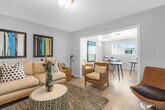 Located at 9651 Graham Street in Cypress, California, this single-floor space offers a thoughtfully arranged layout comprising a living room and a dining room. Despite its compact footprint, the space is designed to maximize functionality and comfort.

The living room features light wood-style flooring, neutral painted walls, and a wall-mounted flat-screen television. A plush sofa anchored by a round patterned coffee table sits atop a textured area rug, while wicker accent chairs and side tables add a warm, natural element. Framed artwork in teal and black tones provides a cohesive decorative touch, and a glass-panel exterior door invites natural light from outside.

The dining room is bright and airy, benefiting from multiple windows dressed with vertical blinds that allow generous natural light to filter in. The space accommodates several rustic wood tables with mid-century modern hairpin-style legs paired with contemporary white chairs, making it well-suited for both dining and collaborative use.

The two areas connect seamlessly through a wide, white-trimmed archway, creating an open and cohesive flow throughout the single floor. Clean lines, consistent light wood flooring, and a soft neutral palette throughout lend the space a calm, welcoming ambiance well-suited for a variety of uses.