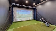 Located at 632 Newark Avenue in Jersey City, NJ, this purpose-built indoor golf simulator bay offers a fully immersive golfing experience in a compact, well-appointed space. The room is thoughtfully designed with dark charcoal walls and a black ceiling that minimize light interference and enhance the visual impact of the large projection screen at the front of the bay. A TruGolf simulator system powers the experience, displaying detailed, lifelike course environments on the expansive screen for a realistic round of golf regardless of weather or season. The hitting area features artificial turf flooring with dedicated hitting mats, providing an authentic feel underfoot during play. A side netting barrier runs along the wall to safely contain errant shots. Recessed ceiling lighting illuminates the space efficiently without compromising the simulator display. Adjacent to the hitting area, a lounge zone with stylish striped pod chairs and a central control station offers a comfortable spot for players to relax between shots and monitor swing data. The space transitions seamlessly between active play and social lounging, making it suitable for solo practice sessions or group outings. The overall ambiance is modern, focused, and sport-oriented, delivering a premium golf simulation experience in the heart of Jersey City.