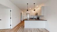 This contemporary single-floor space is thoughtfully designed with a clean, modern aesthetic throughout. The kitchen serves as a central highlight, featuring a peninsula with a dark stone countertop, white upper cabinetry, gray lower cabinetry, and a classic subway tile backsplash. Stainless steel appliances — including a French door refrigerator, electric range, and over-the-range microwave — complete the functional and stylish setup. Pendant lighting above the peninsula adds a warm, refined touch alongside recessed ceiling lights.

Light wood-style flooring flows seamlessly throughout the main living areas and bedrooms, creating a bright and cohesive feel. The bedrooms are well-sized, each featuring windows that allow natural light to fill the space, roller shades for privacy, and individual climate control units.

The unit includes two bathrooms. The full bathroom offers a white vanity with under-cabinet storage, a backlit mirror, and a glass-enclosed walk-in shower with tile detailing. The second bathroom is more utilitarian, finished with large-format neutral tiles and accessibility grab bars. A hallway connects the living and sleeping areas, maintaining a logical and efficient layout throughout the space.