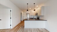 This contemporary single-floor space is thoughtfully designed with a clean, modern aesthetic throughout. The kitchen serves as a central highlight, featuring a peninsula with a dark stone countertop, white upper cabinetry, gray lower cabinetry, and a classic subway tile backsplash. Stainless steel appliances — including a French door refrigerator, electric range, and over-the-range microwave — complete the functional and stylish setup. Pendant lighting above the peninsula adds a warm, refined touch alongside recessed ceiling lights.

Light wood-style flooring flows seamlessly throughout the main living areas and bedrooms, creating a bright and cohesive feel. The bedrooms are well-sized, each featuring windows that allow natural light to fill the space, roller shades for privacy, and individual climate control units.

The unit includes two bathrooms. The full bathroom offers a white vanity with under-cabinet storage, a backlit mirror, and a glass-enclosed walk-in shower with tile detailing. The second bathroom is more utilitarian, finished with large-format neutral tiles and accessibility grab bars. A hallway connects the living and sleeping areas, maintaining a logical and efficient layout throughout the space.