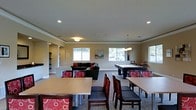 This single-floor community clubhouse is thoughtfully designed to accommodate a variety of social and recreational activities. The open-concept layout allows multiple functional zones to coexist harmoniously, creating a welcoming and versatile gathering space.

The lounge area features a plush dark sofa accented with bold red and black throw pillows, positioned to encourage relaxed conversation. Adjacent to the lounge, a full-size billiards table serves as a central recreational focal point, illuminated by a pendant light fixture that adds a stylish touch overhead.

Throughout the space, multiple dining tables with upholstered chairs — featuring a distinctive red circular pattern — provide ample seating for meals, meetings, or casual gatherings. The mix of seating styles adds visual interest while maintaining a cohesive, comfortable atmosphere.

Natural light flows in through several windows along the perimeter walls, brightening the neutral beige and gray color palette that defines the interior. Recessed ceiling lighting and additional pendant fixtures ensure the space is well-lit at all times. Crown molding along the ceiling adds a refined architectural detail. The carpeted flooring contributes to a warm, inviting ambiance, making this clubhouse an ideal setting for community engagement and leisure.