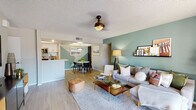 This thoughtfully designed single-floor residence showcases a cohesive blend of modern aesthetics and functional living. The open-concept layout seamlessly connects the living, dining, and kitchen areas, creating a welcoming and airy atmosphere throughout. The living room is anchored by a striking sage-green accent wall adorned with a floating display shelf, complemented by light wood-style flooring, a ceiling fan fixture, and a sliding glass door that opens to a balcony with outdoor views. The dining area features a bold geometric mural in muted teal tones, a round marble-top table, and pendant lighting, adding an artistic focal point to the space. The galley-style kitchen is well-appointed with white upper cabinetry, warm wood lower cabinets, stainless steel appliances, a hexagon-tile backsplash, and a skylight detail above, maximizing both style and utility. The primary bedroom features a dramatic dark accent wall, light wood flooring, and ample natural light from vertical blinds. The second bedroom is bright and versatile with large windows. The bathroom includes a rich dark wood vanity, round mirror, and modern lighting, conveniently situated alongside an in-unit laundry area with side-by-side washer and dryer. Every detail reflects a deliberate, design-forward sensibility.