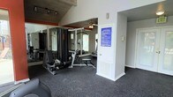 This fitness center offers a practical and functional workout environment designed to meet a variety of exercise needs. The space is outfitted with a multi-station strength training machine, ideal for targeting multiple muscle groups, alongside cardio equipment to support a well-rounded fitness routine. Full-length mirrored walls line one side of the room, enhancing the sense of space while allowing users to monitor their form during exercise. The flooring is covered with durable dark rubber gym matting, providing both safety and comfort during workouts. A ceiling fan overhead helps maintain air circulation throughout the space. Track lighting is mounted above, ensuring the area is well-lit during use. A posted fitness rules sign near the entrance keeps guidelines visible and accessible to all users. The single-floor layout is straightforward and easy to navigate, with a dedicated entryway that separates the fitness area from adjacent spaces. The overall ambiance is focused and purposeful, with an interior design that prioritizes utility and functionality, making it a convenient and effective facility for residents or members seeking an on-site exercise option.