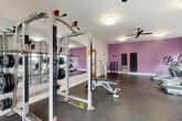 Located at 2401 Clubhouse Ct in Harrisonburg, Virginia, this fitness center offers a well-equipped, single-floor workout facility designed to support a full range of exercise routines. The space features durable rubber flooring throughout, providing a safe and practical surface for both cardio and strength training activities. A row of TRUE-brand treadmills and elliptical machines lines one side of the room, complemented by a rowing machine for varied cardio options. The strength training area includes a multi-function cable and power rack machine paired with an adjustable weight bench, as well as a three-tier dumbbell rack stocked with a comprehensive set of free weights. Large windows along the exterior walls allow generous natural light to fill the room while offering views of the surrounding community. Ceiling fans and flush-mount light fixtures ensure consistent airflow and illumination. Full-length wall mirrors on one side of the room enhance the sense of space and assist with form during workouts. A bold purple accent wall adds a energizing visual element to the interior. The facility also includes a water fountain, wall-mounted televisions, and a dedicated storage shelf unit. A glass-panel entry door provides convenient exterior access, rounding out this practical and well-appointed fitness space.