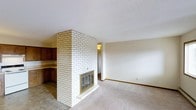 This single-floor space is defined by a striking white-painted brick fireplace that serves as the architectural centerpiece, rising from floor to ceiling and dividing the living area from the adjacent kitchen zone. The fireplace features a classic brass-framed glass door insert, adding a traditional yet functional detail to the room. The living area is finished with neutral wall-to-wall carpeting that creates a warm, understated atmosphere, while the kitchen and transitional areas feature tile flooring, providing a practical contrast. The kitchen, visible to the left of the fireplace, includes dark wood cabinetry with an open shelving section and a countertop bar area, offering a casual and functional layout. Walls throughout are painted in soft, neutral tones, contributing to a clean and open feel. A textured ceiling adds subtle dimension to the space. The overall design is straightforward and practical, with the white brick fireplace lending a distinctive character to what is otherwise a simple, livable layout suited to everyday comfort.