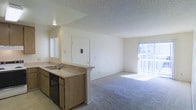 This single-floor unit features an open-concept design that seamlessly connects the kitchen and living area, creating a spacious and functional layout. The kitchen is positioned along one side and includes a double-basin sink, a dishwasher, wood-toned cabinetry, and a countertop that extends into a breakfast bar, providing a natural boundary between the kitchen and the main living space. A mirror above the sink adds visual depth to the area.

The living area is finished with wall-to-wall carpet, offering a comfortable and cohesive feel throughout. A sliding glass door along the far wall floods the room with natural light and provides direct access to an outdoor area, enhancing the sense of openness. Vertical blinds offer privacy and light control as needed.

The front entry door features a keypad access panel, adding a layer of convenience and security. The unit's ceilings are finished with a textured popcorn treatment and include a centrally mounted light fixture. The overall color palette is neutral, with white walls and light-toned surfaces contributing to a clean, bright interior ambiance well-suited for everyday living.