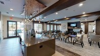 Located at 5173 Forest Lane Place in Dallas, Texas, this thoughtfully designed single-floor communal space blends contemporary elegance with functional versatility. The layout encompasses distinct zones that flow seamlessly into one another, creating a welcoming and refined environment throughout.

The dining area serves as a focal point, anchored by a striking central island with a polished countertop beneath a dramatic cluster of Edison bulb pendant lights suspended from a raw-edge wood accent piece. Dark exposed wood ceiling beams add warmth and architectural character. Multiple dining tables with upholstered seating are arranged beneath tall windows dressed in sheer layered curtains, allowing abundant natural light to fill the space. A stone fireplace with a wall-mounted television provides a cozy gathering point on one end.

A dedicated lounge area features plush, curated furnishings including a sputnik chandelier, bold accent consoles, and patterned armchairs arranged around low tables—ideal for conversation and relaxation. A separate, quieter sitting room offers a more intimate layout with neutral-toned sofas, accent chairs, and floor lamps flanking large windows with outdoor views.

The kitchen zone includes dark cabinetry, stainless steel appliances, a wall oven, and a diagonal wood-paneled accent wall with a mounted television, combining practicality with design flair.