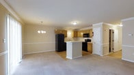 This two-floor residential space offers a well-designed open-concept layout that seamlessly connects the kitchen and living areas. The kitchen is equipped with warm honey-toned cabinetry, black appliances including a refrigerator, microwave, and range, and a white peninsula island that provides additional counter space and serves as a natural divider between the kitchen and the living area. A double sink is positioned along the kitchen counter, complemented by ample cabinet storage above and below. The flooring in the kitchen transitions from hardwood-style to plush neutral carpet in the living area, adding warmth and comfort underfoot. The living space is highlighted by an elegant chandelier with a classic multi-bulb design, lending a touch of sophistication to the room. Neutral-toned walls and white baseboards and trim throughout create a clean, cohesive aesthetic. Recessed lighting and flush-mount ceiling fixtures ensure the space is well-lit. The layout across two floors provides versatile living arrangements, making this a practical and inviting residence suited for a variety of lifestyles.