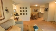 This single-floor residence offers a cohesive and comfortable layout designed for everyday living. The open-concept living and dining area forms the heart of the home, creating a seamless flow between spaces. The living area is furnished with a plush sofa and a wooden coffee table, accented by a classic table lamp that adds a warm, inviting glow. A decorative wall arrangement of ornamental medallions serves as a distinctive focal point, adding character and visual interest to the space. Hardwood-style flooring runs throughout, lending a clean, unified aesthetic that ties the rooms together. The dining area accommodates a full wooden table with seating for multiple guests, positioned beneath soft ceiling lighting. Framed artwork on the walls adds a refined touch to the dining space. Just beyond, a kitchen counter pass-through is visible, hinting at a connected kitchen layout that supports both casual meals and entertaining. Neutral warm tones on the walls complement the natural wood finishes of the furniture and flooring, creating an overall ambiance that is both relaxed and polished. The thoughtful arrangement of the space maximizes functionality while maintaining an open, airy feel throughout.