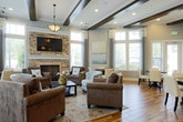 Located at 4285 Johns Creek Pkwy in Suwanee, Georgia, this single-story clubhouse offers a thoughtfully designed gathering space that blends classic architectural details with comfortable, inviting interiors. The main lounge serves as the centerpiece of the space, anchored by a floor-to-ceiling stacked stone fireplace with a mounted flat-screen television above. Dark wood ceiling beams add warmth and definition to the room's generous height, while recessed lighting and a pendant fixture provide layered illumination throughout. A patterned area rug in soft, earthy tones anchors an arrangement of plush upholstered sofas and accent chairs in neutral browns and muted blues, creating a relaxed yet polished atmosphere. Large windows dressed with flowing curtains allow natural light to fill the room and offer views of the surrounding outdoor greenery. Adjacent dining areas feature dark round tables paired with cream upholstered chairs, positioned near expansive windows that further enhance the connection to the outdoors. Wood-look flooring flows throughout the space, complementing the warm color palette. A kitchenette area with dark cabinetry, granite countertops, a stacked stone accent wall, and a utility sink provides added functionality for events and gatherings. Together, these elements create a refined yet approachable community space.