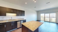 Located at 2645 Railroad Street in Pittsburgh's dynamic Strip District, this well-appointed one-bedroom, one-bathroom residence offers a thoughtfully designed open-concept floor plan. The combined kitchen and living area serves as the heart of the home, showcasing dark espresso cabinetry that extends to the ceiling, granite countertops, and a full suite of stainless steel appliances including a refrigerator, range, microwave, and dishwasher. A central kitchen island with a matching granite surface provides additional prep space and casual dining potential. Rich hardwood-style flooring runs throughout, adding warmth and continuity to the space. The living area flows naturally toward a sliding glass door that opens to a private balcony, offering elevated views of the Pittsburgh skyline and surrounding greenery. The bedroom is generously sized, featuring a large window with outdoor views, a curtain rod for window treatments, and ample natural light. Three closets provide excellent storage throughout the unit. The bathroom features a granite-topped dark vanity, consistent with the home's cohesive design language. Additional conveniences include in-unit laundry and a hallway that connects all areas efficiently. The overall aesthetic is clean, modern, and move-in ready.