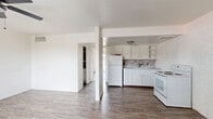 This single-floor unit offers a practical and straightforward layout encompassing a combined kitchen and living area alongside one bedroom. The interior is finished in a bright, all-white color scheme throughout, with wood-look plank flooring running continuously across every room, lending a cohesive and clean aesthetic.

The open living area greets you at the entrance, featuring a white paneled front door and a window fitted with blinds that allows natural light to filter in. A painted white brick accent wall adds a subtle textural element to the space. A ceiling fan with a light fixture provides both air circulation and overhead lighting.

The kitchen is positioned adjacent to the living area and is defined by white cabinetry with open shelving, a mosaic tile backsplash, a white refrigerator, an electric range, and a single-basin sink. The kitchen also showcases an exposed painted brick wall, adding character to the functional space.

The spacious primary bedroom offers generous room, featuring three interior doors — providing access to closets and additional storage — along with a wall-mounted HVAC unit for climate control.

Throughout the unit, the consistent use of white walls, white trim, and wood-look flooring creates a unified and easy-to-style interior.