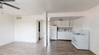 This single-floor unit offers a practical and straightforward layout encompassing a combined kitchen and living area alongside one bedroom. The interior is finished in a bright, all-white color scheme throughout, with wood-look plank flooring running continuously across every room, lending a cohesive and clean aesthetic.

The open living area greets you at the entrance, featuring a white paneled front door and a window fitted with blinds that allows natural light to filter in. A painted white brick accent wall adds a subtle textural element to the space. A ceiling fan with a light fixture provides both air circulation and overhead lighting.

The kitchen is positioned adjacent to the living area and is defined by white cabinetry with open shelving, a mosaic tile backsplash, a white refrigerator, an electric range, and a single-basin sink. The kitchen also showcases an exposed painted brick wall, adding character to the functional space.

The spacious primary bedroom offers generous room, featuring three interior doors — providing access to closets and additional storage — along with a wall-mounted HVAC unit for climate control.

Throughout the unit, the consistent use of white walls, white trim, and wood-look flooring creates a unified and easy-to-style interior.