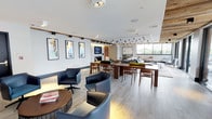 This contemporary single-floor space is thoughtfully designed to balance functionality with refined aesthetics. The open-plan layout creates a welcoming, airy atmosphere enhanced by large windows that flood the interior with natural light and offer views of the surrounding greenery. Light wood-toned flooring runs throughout, contributing to the bright and clean visual character of the space.

The lounge area is furnished with a mix of comfortable navy blue leather bucket chairs and swivel seats, arranged around small accent tables to encourage casual conversation or individual use. A series of long wooden communal tables with matching mid-century-style chairs provide ample space for group gatherings, dining, or collaborative work.

Overhead, a combination of recessed lighting, sleek track lighting, and a striking pendant chandelier with downward-facing bulbs adds layered illumination that enhances the space's modern ambiance. The walls are adorned with a curated selection of framed artwork in bold, vibrant hues, adding personality and visual interest throughout.

Toward the far end, a soft seating lounge area with sofas and additional chairs creates a more relaxed setting, ideal for unwinding. A wall-mounted television and fireplace surround anchor this zone, adding both comfort and function. Overall, the space presents a polished, modern environment suited for a variety of social and professional uses.