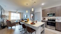 This single-floor residence showcases a thoughtfully designed open-concept layout that integrates the kitchen, dining, and living areas into one cohesive, flowing space. The kitchen is a standout feature, anchored by a large central island with a crisp white quartz-style countertop and seating for multiple guests via sleek brown leather bar stools. Pendant lighting hangs above the island, adding a warm, modern aesthetic. Cabinetry finished in rich wood tones lines the kitchen walls, complemented by stainless steel appliances and ample counter space.

The living area extends naturally from the kitchen, bathed in natural light from large windows dressed with curtain panels. A ceiling fan overhead provides both function and style, while the open floor plan creates a generous sense of space. Wood-style flooring runs throughout, tying the areas together cohesively.

A hallway visible in the background leads to additional rooms, including what appears to be a bedroom, hinting at a well-organized floor plan. The overall ambiance is clean, contemporary, and welcoming, with neutral tones and modern finishes throughout.