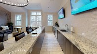 This well-appointed clubhouse offers a welcoming, open-concept layout designed for both casual gatherings and community events. Light wood-look tile flooring runs throughout, complementing the neutral warm tones of the walls and creating a bright, cohesive aesthetic.

The centerpiece of the space is a generously sized granite-topped bar with integrated sink, flanked by stainless steel refrigerators and backed by a textured penny-tile wall. A row of upholstered bar stools invites guests to gather, while dual wall-mounted flat-screen TVs provide entertainment. A dramatic circular chandelier with exposed bulbs anchors the bar area and adds a modern, social atmosphere.

Adjacent to the bar, a full kitchenette features additional granite countertops, dark cabinetry, and a wall-mounted TV — all bathed in natural light from large double-hung windows that frame views of surrounding trees. This area also includes additional seating, making it a versatile space for events or catered functions.

The lounge area at the far end of the clubhouse is distinguished by a striking large-scale vintage map mural serving as a dramatic accent wall, evoking a sense of local pride and character. Plush dark sofas with decorative pillows, round wooden bistro tables, and framed black-and-white photography complete the inviting, relaxed atmosphere. Vaulted ceilings throughout enhance the sense of spaciousness and airiness.