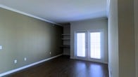 This single-floor space offers a clean and functional layout with thoughtful architectural details throughout. The main living area is finished with rich, dark hardwood flooring that adds warmth and elegance underfoot, complemented by crisp white crown molding that frames the ceiling and white baseboards that provide a polished contrast to the earthy, olive-toned walls.

A standout feature of the space is the built-in shelving unit nestled into a dedicated wall niche, complete with a lower built-in desk surface — an ideal combination for display, storage, and a functional workspace. The shelves are well-proportioned and offer versatile use for books, décor, or everyday essentials.

Natural light flows generously into the room through a set of classic French doors, fitted with horizontal blinds for privacy and light control. The doors offer both aesthetic appeal and practical access, brightening the interior with an airy, open feel.

The overall ambiance is calm and understated, with neutral tones and traditional design elements creating a welcoming atmosphere. The space is move-in ready and well-suited for a variety of living arrangements, balancing style and practicality in a cohesive, single-level layout.