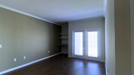 This single-floor space offers a clean and functional layout with thoughtful architectural details throughout. The main living area is finished with rich, dark hardwood flooring that adds warmth and elegance underfoot, complemented by crisp white crown molding that frames the ceiling and white baseboards that provide a polished contrast to the earthy, olive-toned walls.

A standout feature of the space is the built-in shelving unit nestled into a dedicated wall niche, complete with a lower built-in desk surface — an ideal combination for display, storage, and a functional workspace. The shelves are well-proportioned and offer versatile use for books, décor, or everyday essentials.

Natural light flows generously into the room through a set of classic French doors, fitted with horizontal blinds for privacy and light control. The doors offer both aesthetic appeal and practical access, brightening the interior with an airy, open feel.

The overall ambiance is calm and understated, with neutral tones and traditional design elements creating a welcoming atmosphere. The space is move-in ready and well-suited for a variety of living arrangements, balancing style and practicality in a cohesive, single-level layout.