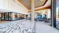 Located at 1873 N Rollins Rd in Burlingame, California, this expansive ground-floor lobby spans an impressive footprint across 10 distinct areas, delivering a refined and welcoming first impression. The space is defined by soaring double-height ceilings, intricate patterned marble tile flooring, and floor-to-ceiling glass curtain walls that draw in abundant natural light and offer open views of the surrounding landscape.

The main entry foyer features a striking geometric mosaic tile floor that anchors the space, complemented by bold teal architectural columns and sculptural décor pieces. Multiple lounge areas are thoughtfully arranged throughout, including a curved rose-pink banquette seating nook beneath a cascading globe chandelier, intimate clusters of plush barrel chairs, and a long communal table with elegant brass-accented pendant lighting above.

A standout large-scale abstract painting in blues and teals commands one wall, adding a vibrant artistic focal point. The elevator vestibule is outfitted with mirrored ceilings, warm Edison-style pendant lights, and richly tiled surrounds. Warm wood-toned slat ceilings with recessed gold-accent lighting transition beautifully between zones, while a glass-enclosed reception structure anchors the interior. The overall ambiance is contemporary, sophisticated, and thoughtfully curated throughout.