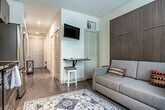 Located at 1817 E Washington Ave in Madison, Wisconsin, this efficiently designed single-floor unit offers a thoughtfully arranged layout comprising a kitchen, hallway, and bathroom. The hallway serves as the spine of the space, finished with smooth hardwood-style flooring and crisp white walls illuminated by recessed lighting, creating a clean and welcoming passage. A window at the far end draws in natural light, enhancing the sense of depth and openness.

The kitchen is a standout feature, outfitted with rich dark wood cabinetry extending to the ceiling for maximum storage, granite countertops, and a full suite of stainless steel appliances including a refrigerator, range, microwave, and dishwasher. A window beside the counters allows natural light to brighten the workspace. The bathroom is finished with stone-look tile flooring, a wood-toned vanity with granite countertop, a large mirror, and a walk-in shower, offering a clean and functional design.

The building itself features a well-appointed courtyard with seating areas, a pergola, and lush landscaping — providing residents with attractive outdoor communal space. The overall aesthetic throughout the unit is modern and cohesive, combining warm wood tones with neutral finishes for a comfortable, contemporary feel.