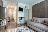 Located at 1817 E Washington Ave in Madison, Wisconsin, this efficiently designed single-floor unit offers a thoughtfully arranged layout comprising a kitchen, hallway, and bathroom. The hallway serves as the spine of the space, finished with smooth hardwood-style flooring and crisp white walls illuminated by recessed lighting, creating a clean and welcoming passage. A window at the far end draws in natural light, enhancing the sense of depth and openness.

The kitchen is a standout feature, outfitted with rich dark wood cabinetry extending to the ceiling for maximum storage, granite countertops, and a full suite of stainless steel appliances including a refrigerator, range, microwave, and dishwasher. A window beside the counters allows natural light to brighten the workspace. The bathroom is finished with stone-look tile flooring, a wood-toned vanity with granite countertop, a large mirror, and a walk-in shower, offering a clean and functional design.

The building itself features a well-appointed courtyard with seating areas, a pergola, and lush landscaping — providing residents with attractive outdoor communal space. The overall aesthetic throughout the unit is modern and cohesive, combining warm wood tones with neutral finishes for a comfortable, contemporary feel.