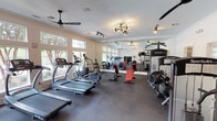 This fitness center offers a comprehensive workout environment spread across a single level, thoughtfully designed to accommodate a wide range of exercise needs. The main cardio area is lined with large windows that flood the space with natural light and provide refreshing views of mature trees and an outdoor pool, creating an inviting atmosphere for exercise. SportsArt treadmills and elliptical machines are arranged along the windowed walls, complemented by ceiling fans that promote air circulation throughout. A full-wall mirror visually expands the space and allows members to monitor their form during workouts. The strength training section is equipped with a multi-functional cable machine, a lat pulldown station, adjustable benches, and a dumbbell rack, catering to a full range of resistance training exercises. A dedicated studio area features a spin bike, a rowing machine, wall-mounted exercise mats, and a flat-screen television, making it ideal for cycling sessions or guided workouts. Track lighting and a stylish sputnik chandelier provide focused illumination throughout the facility. The dark rubber flooring throughout is both durable and practical, while the clean white walls and crown molding lend the space a polished, well-maintained aesthetic.