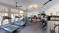 This fitness center offers a comprehensive workout environment spread across a single level, thoughtfully designed to accommodate a wide range of exercise needs. The main cardio area is lined with large windows that flood the space with natural light and provide refreshing views of mature trees and an outdoor pool, creating an inviting atmosphere for exercise. SportsArt treadmills and elliptical machines are arranged along the windowed walls, complemented by ceiling fans that promote air circulation throughout. A full-wall mirror visually expands the space and allows members to monitor their form during workouts. The strength training section is equipped with a multi-functional cable machine, a lat pulldown station, adjustable benches, and a dumbbell rack, catering to a full range of resistance training exercises. A dedicated studio area features a spin bike, a rowing machine, wall-mounted exercise mats, and a flat-screen television, making it ideal for cycling sessions or guided workouts. Track lighting and a stylish sputnik chandelier provide focused illumination throughout the facility. The dark rubber flooring throughout is both durable and practical, while the clean white walls and crown molding lend the space a polished, well-maintained aesthetic.
