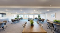 Located at 6039 Seminole Trail in Barboursville, Virginia, this single-floor community space offers a generous and well-appointed environment designed for gathering, collaboration, and socializing. The main hall is expansive and open, accommodating multiple large wooden tables surrounded by modern upholstered chairs in neutral and blue tones. Recessed lighting throughout keeps the space bright and welcoming, while a row of wide windows along the perimeter fills the room with natural light and provides views of the surrounding exterior. Patterned carpet tiles in muted tones add texture and comfort underfoot. Tasteful greenery and potted plants are arranged throughout, lending a fresh, lively ambiance. A wall-mounted flat-screen television anchors one end of the main hall, making the space equally suited for presentations or casual viewing. A private conference room, accessible via glass-paneled doors, offers a more intimate setting with its own wooden table, upholstered seating, and large windows overlooking the outdoors. Adjacent to the main area, a functional kitchenette features dark wood cabinetry, granite-style countertops, light hardwood-style flooring, and a full-size stainless steel refrigerator, providing convenient amenity support for events and gatherings.