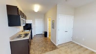This single-floor unit offers a practical and well-organized layout designed for comfortable everyday living. The kitchen is a standout feature, equipped with rich dark espresso cabinetry both above and below the counters, a light-toned laminate countertop, a stainless steel refrigerator, an electric coil cooktop, and a built-in microwave. The kitchen flows into an open entryway area finished with stone-look tile flooring, which provides a durable and easy-to-clean transition space. A set of double doors offers additional storage, while a utility panel is conveniently recessed into the wall nearby. The front entry door is bright white, contributing to a clean, welcoming aesthetic throughout. The bathroom is compact and efficiently arranged, featuring a shower-tub combo, a toilet, and a vanity with dark cabinetry topped with a white sink basin. A lighted mirror cabinet above the vanity adds both function and brightness to the space. The bedroom is finished with plush wall-to-wall carpeting and features a window that invites natural light, fitted with blinds for privacy. A ceiling fan with light fixture provides both comfort and illumination. Neutral warm-toned walls throughout the unit create a cohesive and move-in-ready atmosphere.