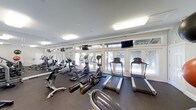 This fitness center offers a clean, modern environment designed to support a well-rounded workout experience. The space is outfitted with a comprehensive selection of cardio equipment, including multiple treadmills, elliptical trainers, and stationary bikes, all arranged to maximize accessibility and comfort. A row of large windows and glass-paneled doors lines one wall, flooding the room with natural light and offering pleasant views of the surrounding greenery, creating an inviting and energizing atmosphere. Wall-mounted flat-screen televisions are positioned throughout the space, ensuring entertainment is always within view during a workout session. Full-length mirrors along one wall enhance the sense of openness while allowing users to monitor their form. The room features a dark, rubberized flooring ideal for fitness use, providing both durability and safety underfoot. Recessed and flush-mounted lighting supplements the natural light, keeping the space well-lit at all times. The overall ambiance is bright, functional, and thoughtfully designed, making this fitness center a practical and welcoming facility for daily exercise.