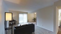 This single-floor residential space is designed with comfort and functionality in mind. The living room serves as the heart of the home, featuring wall-to-wall plush carpeting that adds warmth underfoot and a soft, inviting atmosphere throughout. A large sliding glass door fitted with vertical blinds allows abundant natural light to fill the space while providing access to an outdoor area, creating a seamless indoor-outdoor connection.

The living room is generously sized, comfortably accommodating a full sofa and coffee table arrangement, with ample remaining floor space. Neutral-toned walls keep the aesthetic clean and versatile, complementing a variety of décor styles. The smooth, flat ceiling maintains an uncluttered look across the room.

An open passageway connects the living room to an adjoining room, which features a ceiling fan with light fixture — a practical addition for air circulation and ambient lighting. This secondary space, also carpeted, offers flexibility for use as a dining area, office, or additional sitting room.

Together, the rooms create a cohesive, flowing layout that maximizes usable space. The overall ambiance is bright, neutral, and move-in ready, making this a practical and comfortable single-floor residence suited for a range of lifestyles.