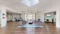 Located at Regal Pointe in Lake Mary, Florida, this single-floor clubhouse is a thoughtfully designed community gathering space that blends modern aesthetics with functional amenities. The centerpiece of the main lounge is a dramatic, oversized skylight that floods the room with natural light, complemented by soaring vaulted ceilings and dual ceiling fans that enhance the airy, open atmosphere. Rich hardwood-style flooring flows throughout, grounding the bright, neutral palette of the white walls and soft gray furnishings.

The main lounge area is generously proportioned, featuring stylishly arranged seating groups — including plush sofas and modern accent chairs set atop a blue wave-patterned area rug — that invite comfortable conversation and relaxation. Floor-to-ceiling drapery-framed sliding glass doors open to views of the outdoor pool area, bringing the Florida sunshine indoors.

A sleek coffee and beverage bar, accented by a textured mosaic tile feature wall and bar-height seating, provides a casual spot for residents to gather. An adjacent business center nook, accessible through a glass-paned French door, offers a quiet workspace equipped with a computer station, mounted display, and comfortable seating. A secondary lounge alcove with a wall-mounted TV rounds out this versatile and welcoming community space.