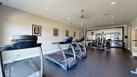 Located at 1061 Spring Creek Dr in Forest, VA, this single-floor fitness and wellness facility is thoughtfully designed to support a full range of health and recovery activities. The main workout area is generously sized, featuring durable rubber flooring, recessed lighting, and ceiling fans that maintain airflow during exercise. Large windows along one wall allow natural light to fill the space and provide views of the surrounding greenery, creating an energizing atmosphere. The cardio zone is equipped with multiple treadmills and an elliptical trainer, while a dedicated strength-training section includes a multi-station cable machine, a dumbbell rack, and a wall-mounted flat-screen television. Full-length mirrors amplify the sense of space and allow users to monitor their form. Motivational framed artwork lines the walls, adding an encouraging aesthetic throughout. Adjacent to the main workout floor, a relaxation area provides a dedicated recovery experience, complete with a massage chair and a private infrared sauna with a warm wood interior. Light tile flooring and soft neutral tones in this area create a calm, spa-like contrast to the active energy of the gym floor. The layout flows efficiently between the exercise and recovery zones, making this facility a comprehensive wellness destination.