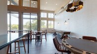 This contemporary amenity lounge is designed to impress, combining stylish industrial elements with warm, inviting finishes. The space is defined by its dramatically soaring double-height ceilings, exposed ductwork, and expansive floor-to-ceiling windows and glass doors that flood the interior with natural light and frame lush tree canopy views beyond an adjacent outdoor balcony. Dark slate-toned tile flooring runs throughout the main gathering area, complementing the neutral warm-beige walls and modern wall sconces.

The main lounge area is furnished with a mix of high-top bistro tables with bar stools, a long glass-top counter with seating, and a dedicated game zone featuring a full-size shuffleboard table. A round poker table surrounded by leather swivel chairs anchors a separate game room nook, accented by a bold geometric pendant light and a wall-mounted flat-screen TV.

A well-appointed wet bar area adds functional elegance, showcasing deep navy-blue cabinetry, a white quartz countertop, a sink, under-counter refrigeration units, and richly textured dark subway tile extending floor to ceiling. A framed Scrabble board adds a playful decorative touch. Together, these zones create a versatile, entertainment-focused space suited for casual gatherings and organized social events alike.